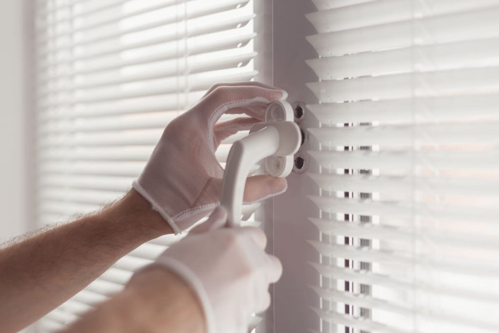 Manos con guantes blancos ajustando persianas en una ventana.